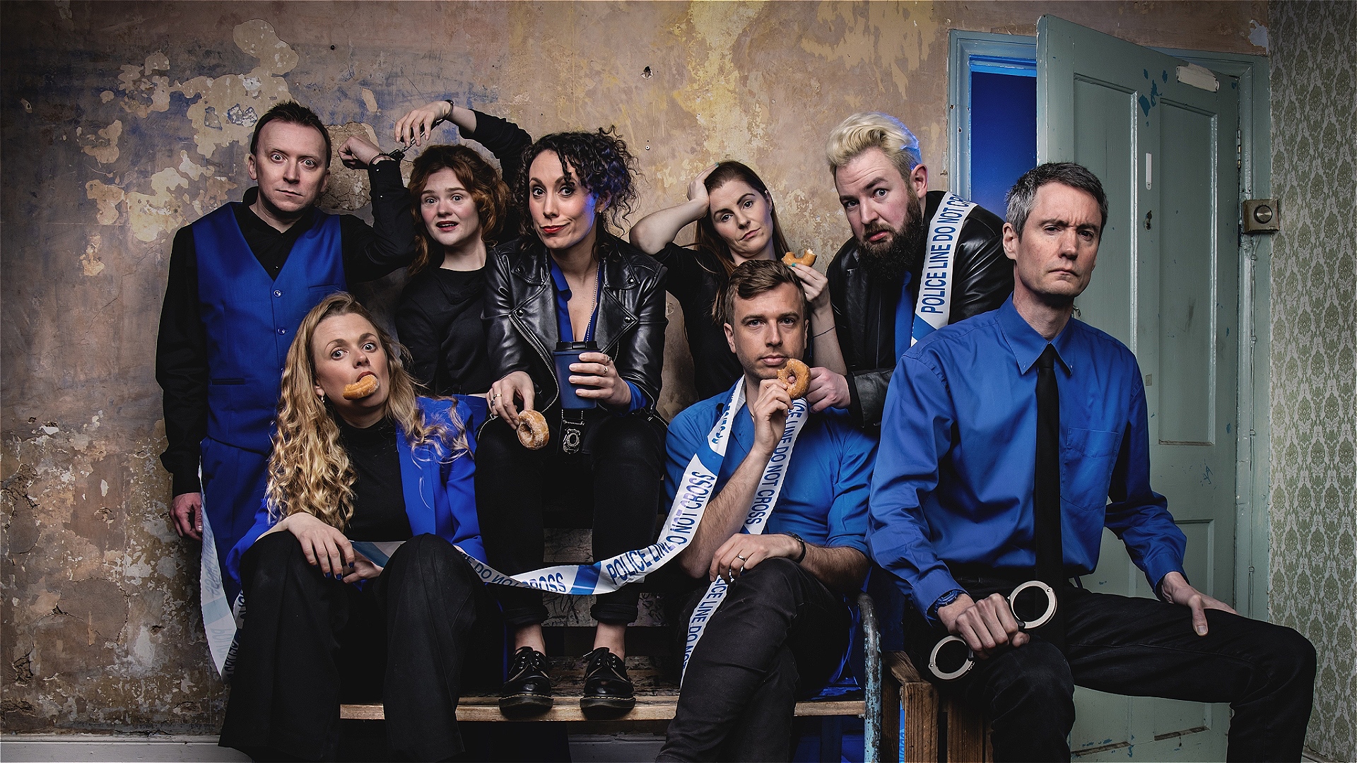 8 actors dressed in blue and black sit on and around a bench. They are comedically handcuffed to each other, eating doughnuts and wrapped in police tape.