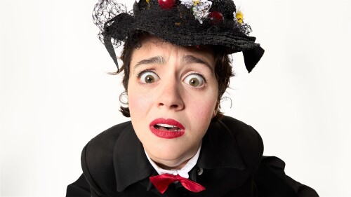 A woman is dressed in a white shirt, red bowtie, black jacket and a black hat with flowers and cherries on them. She is looking into the camera with a confused look, wide eyes and red liptick.