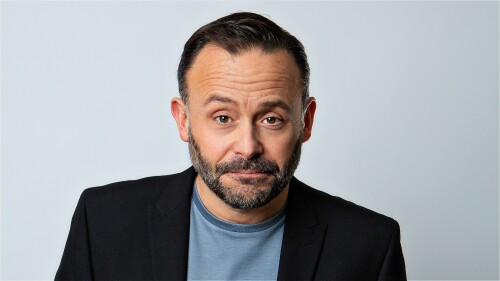 man with a beard and short dark hair wearing a black blazer and blue tshirt in front of a grey background