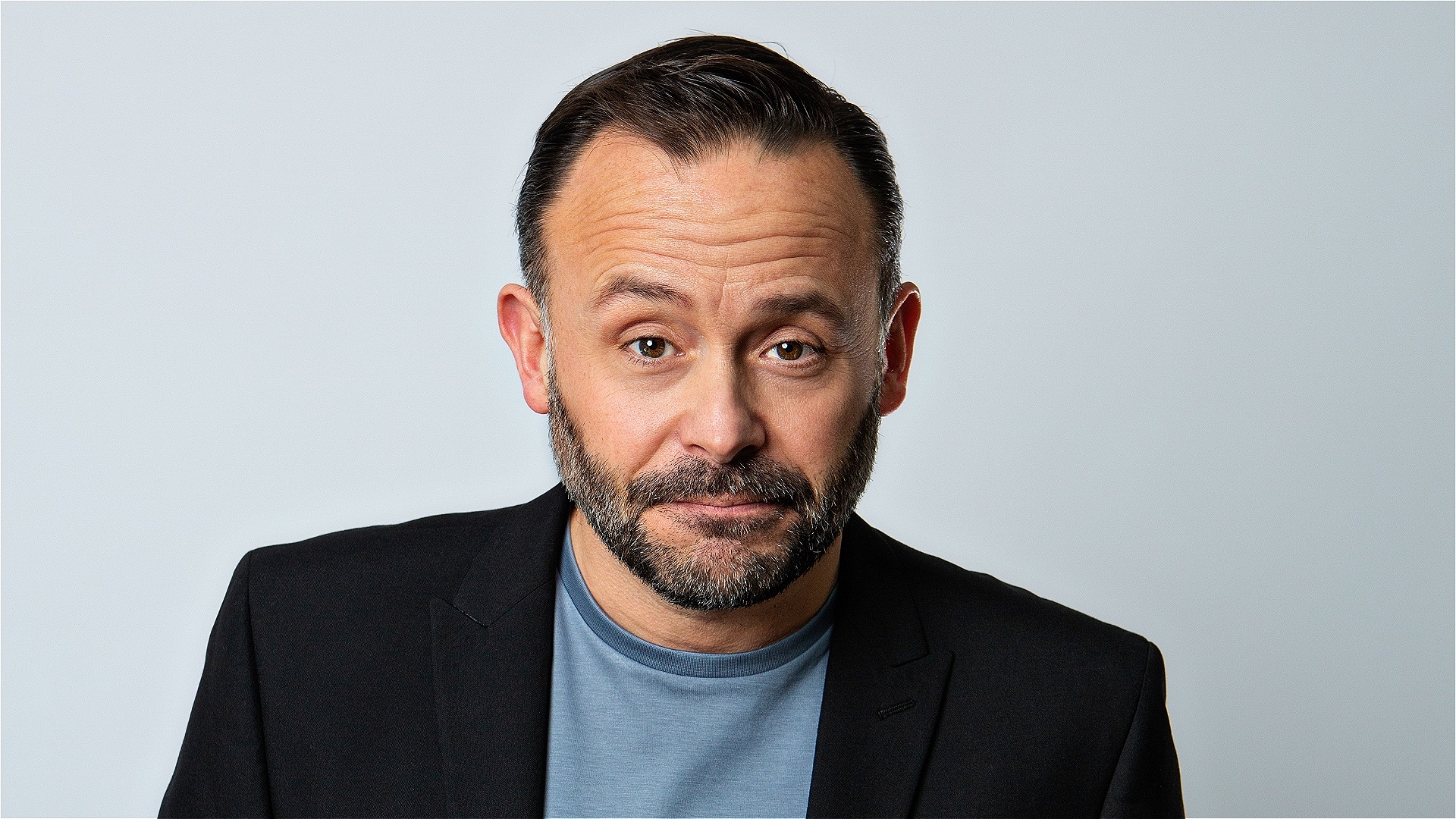 man with a beard and short dark hair wearing a black blazer and blue tshirt in front of a grey background