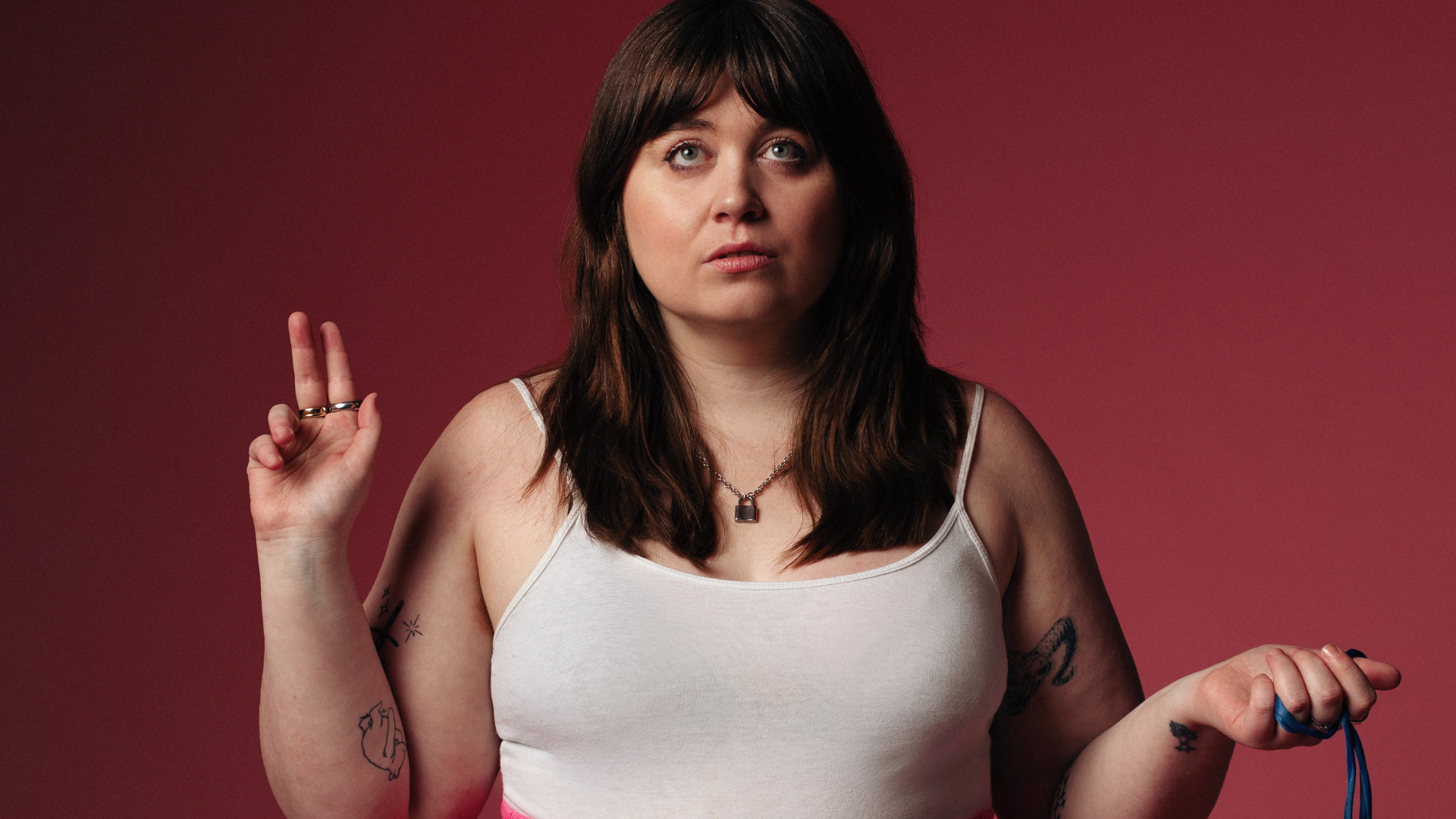 A woman in the centre of the screen is wearing a white vest top and is looking up. Her left hand is pointing up and her right hand is lowered and holding a dark blue ribbon. 