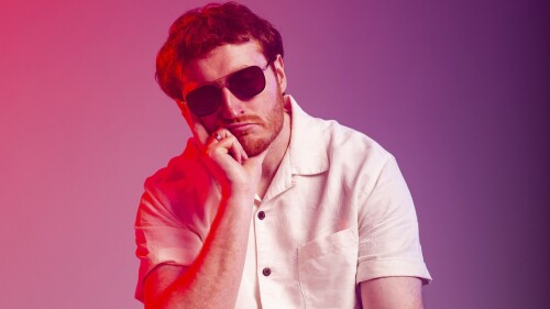 A man wearing sunglasses and a white t-shirt. He is holding his head up with his hand sulkily. 
