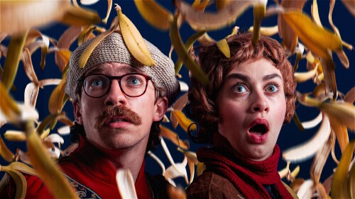 Two people look surprised and fearful as yellow banana peels fall around them against a dark background. One wears glasses and a hat, the other has curly hair and an open mouth.
