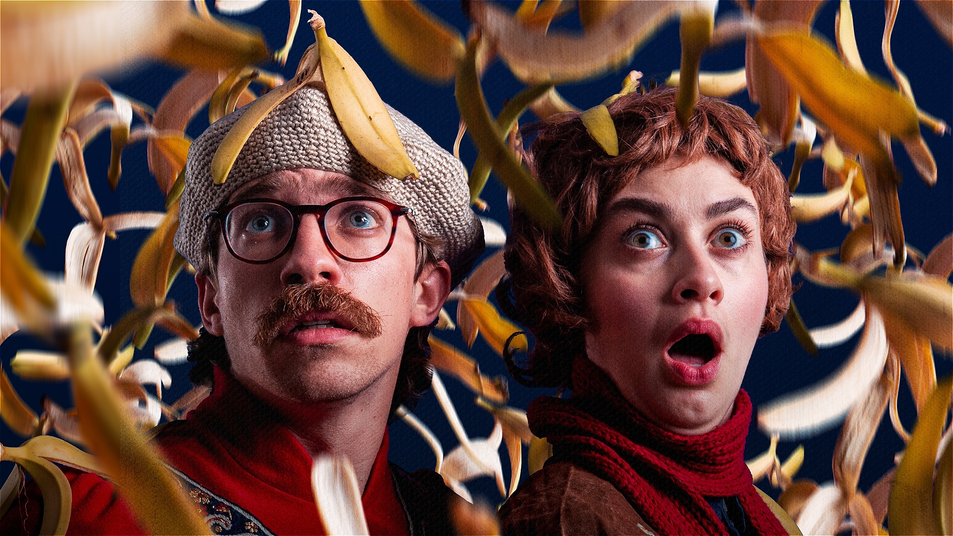 Two people look surprised and fearful as yellow banana peels fall around them against a dark background. One wears glasses and a hat, the other has curly hair and an open mouth.