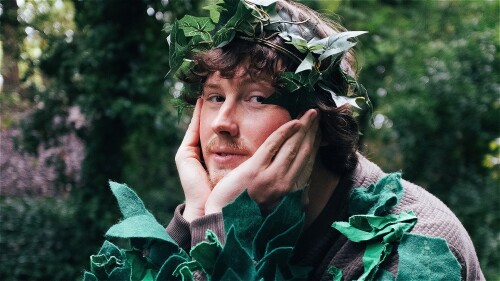 A person dressed in a tree costume made of green felt leaves and a leafy crown poses with their face resting in their hands. They are looking at the camera with a serene expression. The background is a lush, green forest.
