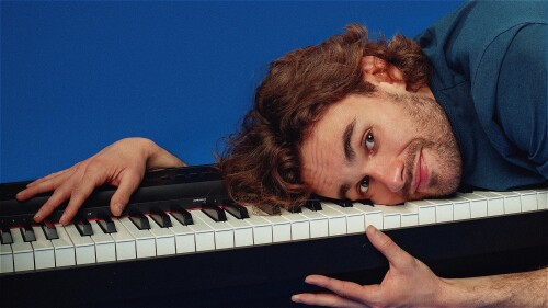 A white man in his early 30s with mid-length curly brown hair and a short brown beard, wearing a blue t-shirt, lies on a piano keyboard with his hands clutching at the sides. He is looking directly at the camera and has a slight smirk on his face.