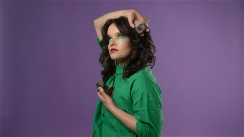 A woman (pronouns she/they) holding a rock over her head. She looks wistful in a green, collared shirt on a lavender backdrop.