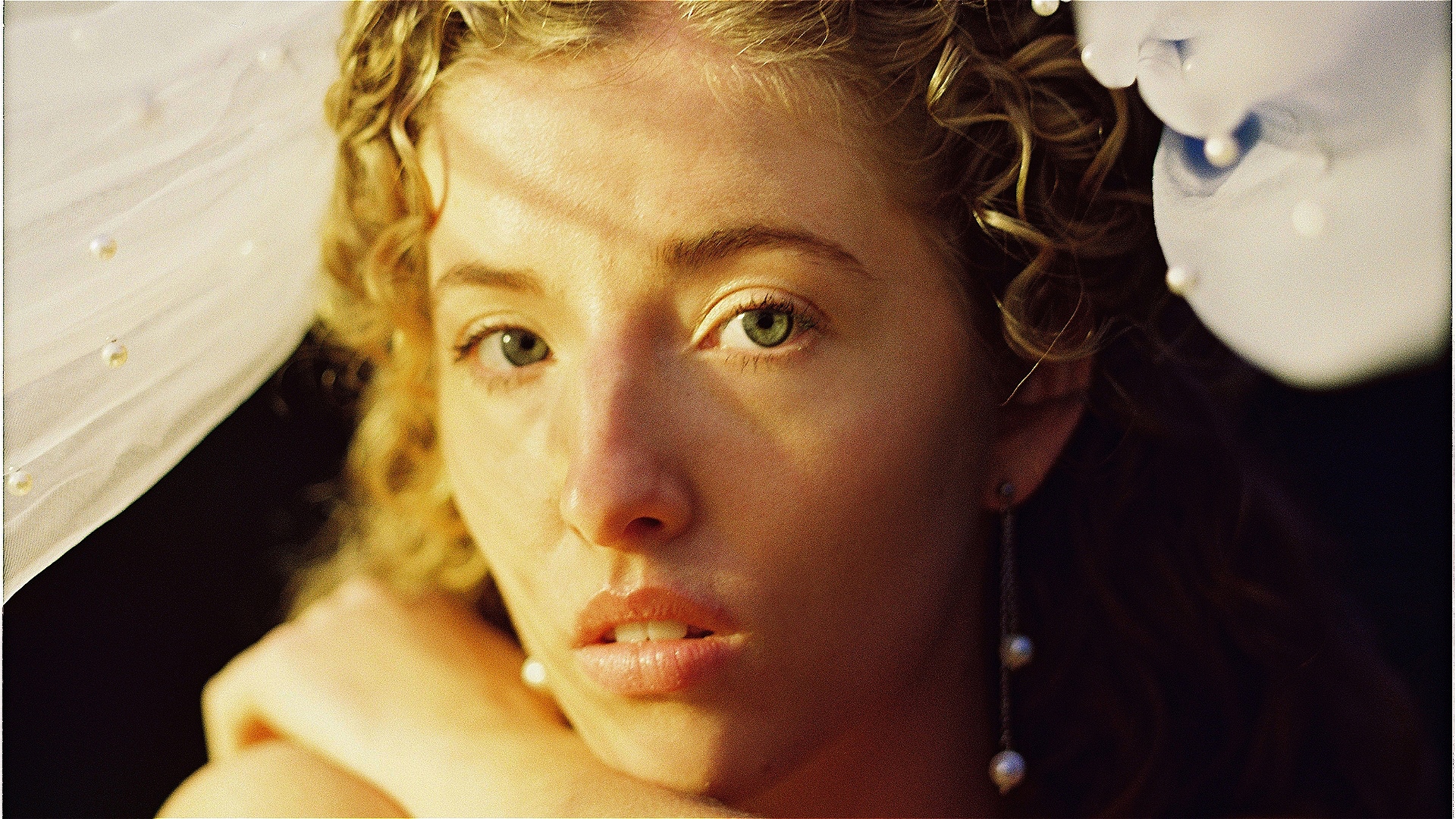 Sutton is hiding under a pearl encrusted veil. Matching pearl earrings dangle from her ears. She is looking straight down the barrel at you. She has green eyes. She looks guarded and concerned, with her lips slightly parted, ready to say something.