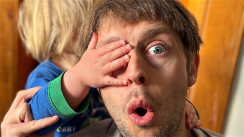 A man looks shocked while a toddler's small hand covers his right eye
