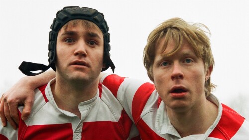 Two white eighteen year old Boys are mid Rugby Scrum. They are both looking forward wearing matching white and red rugby kits. The boy on the left is wearing a black scrum cap and looks nervous, the boy on the right has blonde hair and looks prepared.