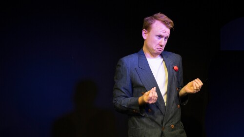 Ben Goldsmith stands on stage wearing a blue suit jacket with a red hankerchief in the top pocket. He is holding his hands out infront of him and clasping them together, he is pulling a sad expression on his face. 