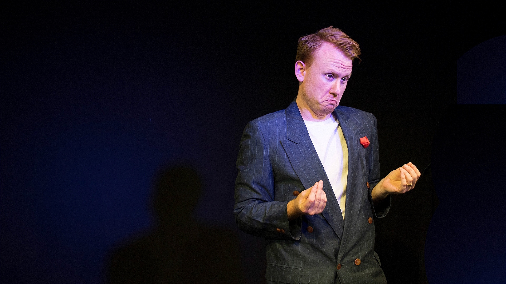 Ben Goldsmith stands on stage wearing a blue suit jacket with a red hankerchief in the top pocket. He is holding his hands out infront of him and clasping them together, he is pulling a sad expression on his face. 