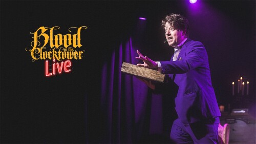 The backdrop is purple curtains and candles with Jon Gracey in a dramatic Storyteller pose in the foreground