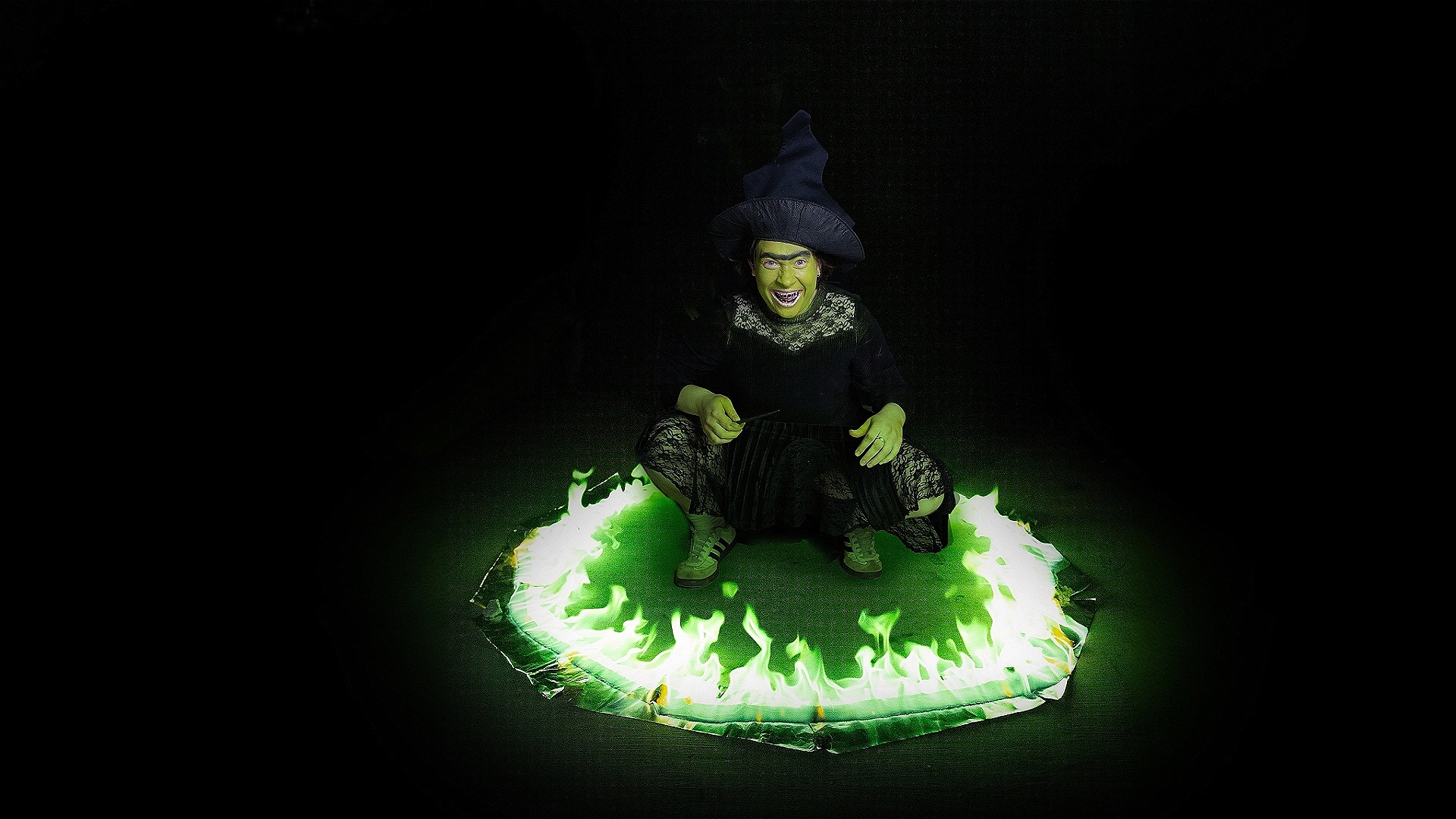 DeliaDelia has a green face and is wearing a witch's hat in full black dress, squatting in the middle of a ring of green fire, holding a wand