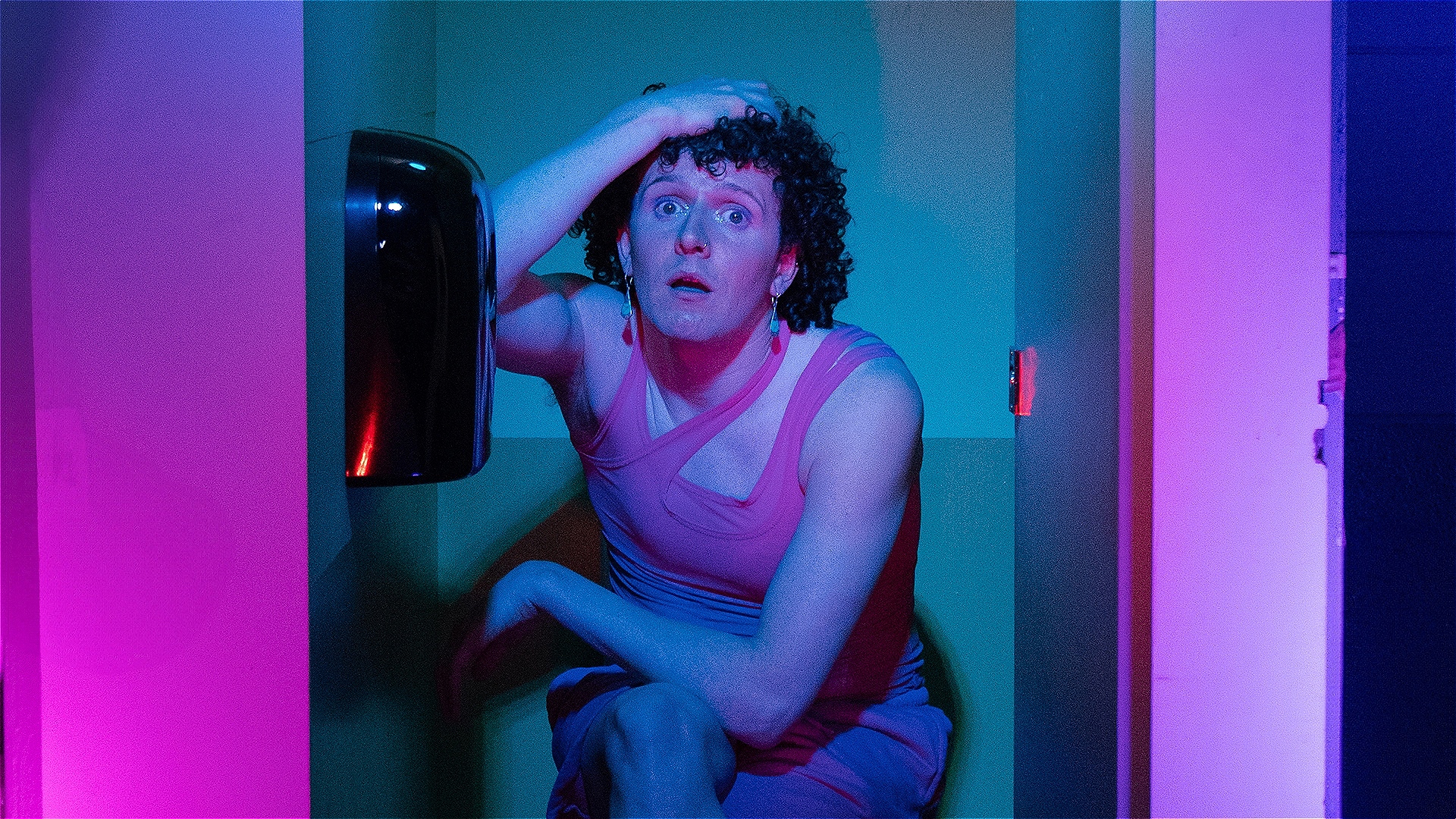 - Three toilet cubicles in colours: pink, green/blue and purple. - Cubicle one (left): A man (Ollie) dangling off the door with a mahoosive smile on his face. - Cubicle two (middle): Ollie sat on the toilet seat with his legs crossed and his hand on his 