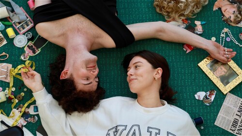 Two friends lie on a green carpet surrounded by cut-outs of Princess Diana and sewing materials. One wears her iconic black revenge dress and holds white rosary beads; the other wears a white YALE jumper and holds a measuring tape. They face each other, 