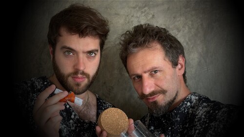 Two white men face the camera in a mysterious way. One holds a rolled up paper tied with an orange bow while the other holds an empty jar with its cap on the other hand.