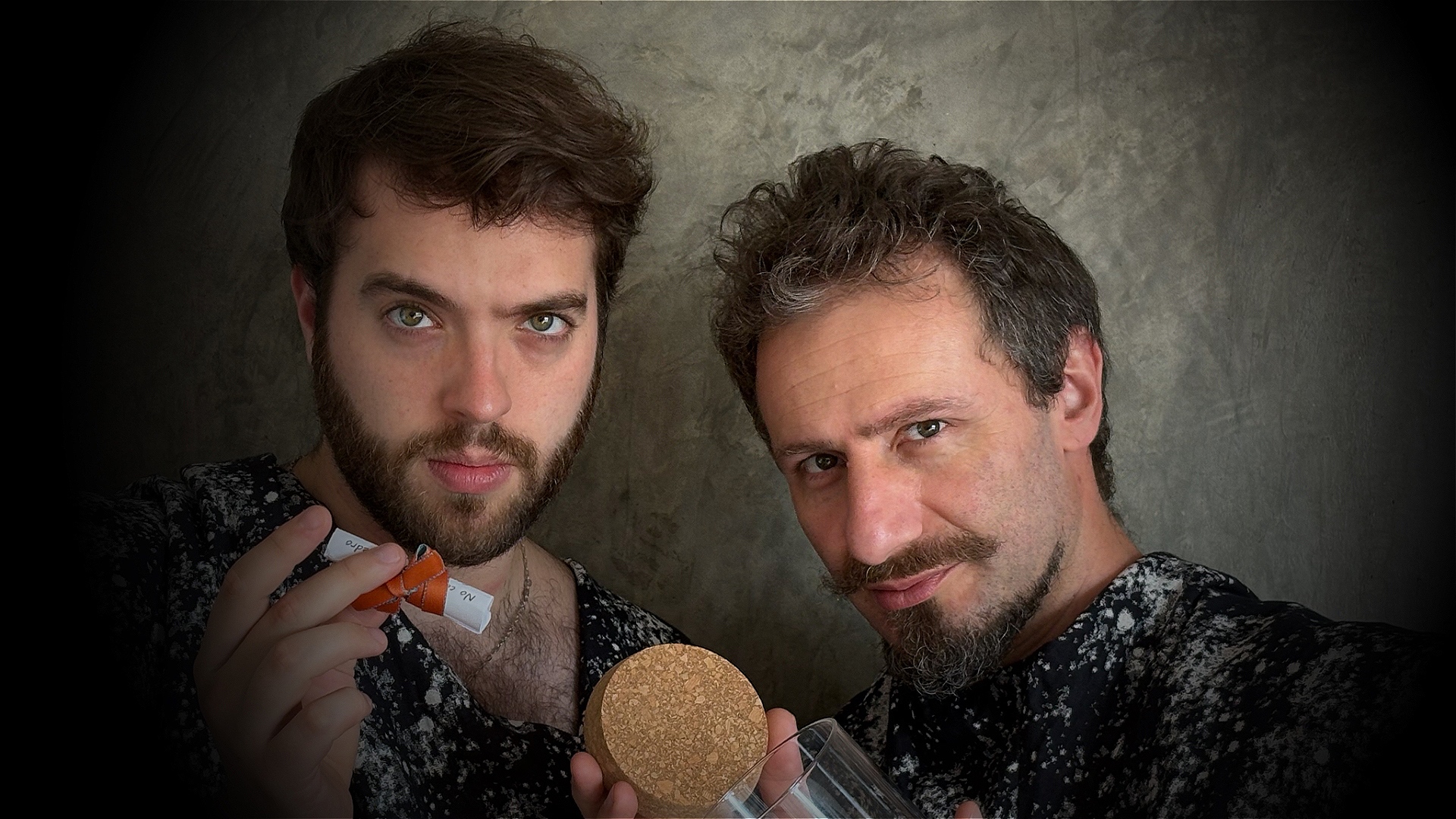 Two white men face the camera in a mysterious way. One holds a rolled up paper tied with an orange bow while the other holds an empty jar with its cap on the other hand.
