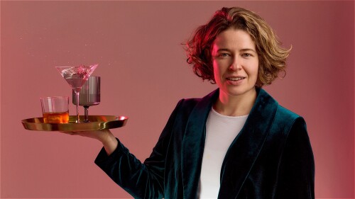 Holly Redford Jones, a white woman with short blonde hair wearing a teal green velvet suit and a white t-shirt, looking straight forward with a smile, and holding in her right hand a gold bar tray with three drinks - a whiskey glass, a martini glass and 