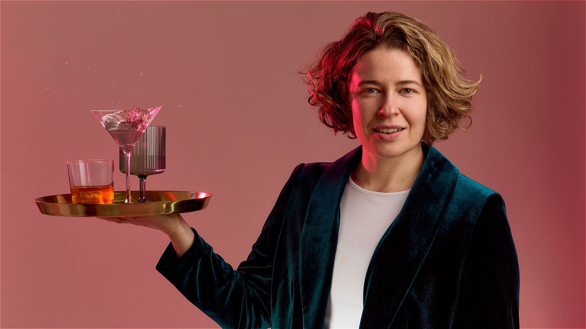 Holly Redford Jones, a white woman with short blonde hair wearing a teal green velvet suit and a white t-shirt, looking straight forward with a smile, and holding in her right hand a gold bar tray with three drinks - a whiskey glass, a martini glass and 