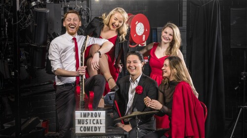 A group of 5 performers holding various musical objects and wearing red and black are laughing and smiling.