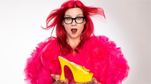 A woman with windswept red hair and black-framed glasses wears a bright pink ruffled dress and holds a daffodil-yellow shoe.