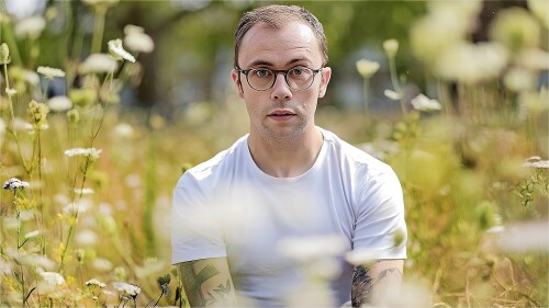 Joe Tracini is sat in a field of wildflowers wearing a white t shirt looking unnecessarily pensive.