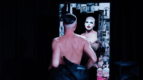 A drag artist in white clown make up stands in front of a mirror that has been adorned with many little photographs and images.