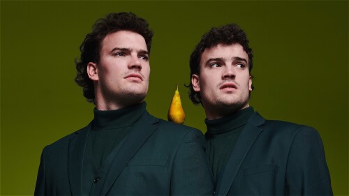 Two twins stand shoulder to shoulder wearing green suits against a similarly green background, a pear rests on their shoulder between them.