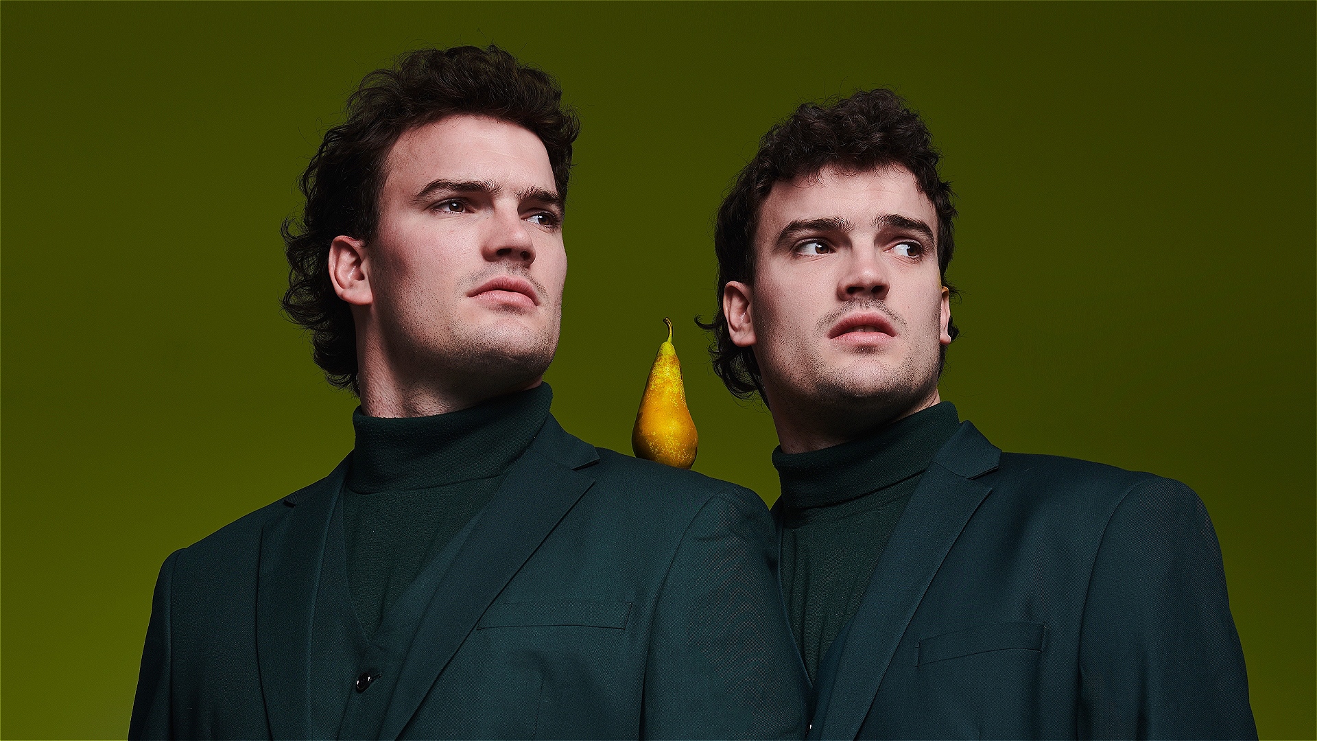 Two twins stand shoulder to shoulder wearing green suits against a similarly green background, a pear rests on their shoulder between them.