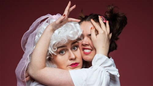 Two women hugging each other. One dressed in a curly wig with a bow and corset. The other dressed in a leotard and ponytails.