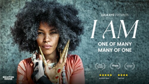 A dark skinned woman (the creator Lula Mebrahtu) is wearing MiMu gloves in one hand and gold jewellery in the other. She is looking directly into the camera as she holds both hands up just under her chin