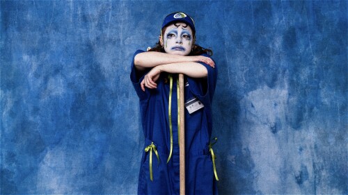 A woman in a blue jumpsuit wearing clown-makeup rests her head on her arms and her forearms on the head of a broom. She is also wearing a blue hat and is stood in front of a blue background.