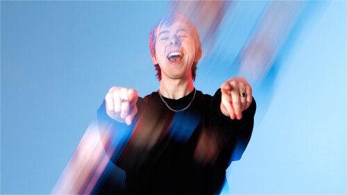 Comedian Chris Turner, wearing a black jersey and silver chain, stands in front of a blue background, pointing both fingers at the camera, moderately distorted by a prism to seem as if he's in motion.