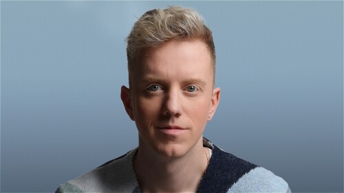 Comedian James Barr in a blue cardigan, close up and looking hopeful, with a slightly shaded face and a light ish blue background.