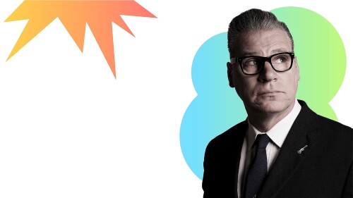 Mark Kermode in the right corner of the screen looking out. He is wearing a black suit and tie and black framed glasses. 