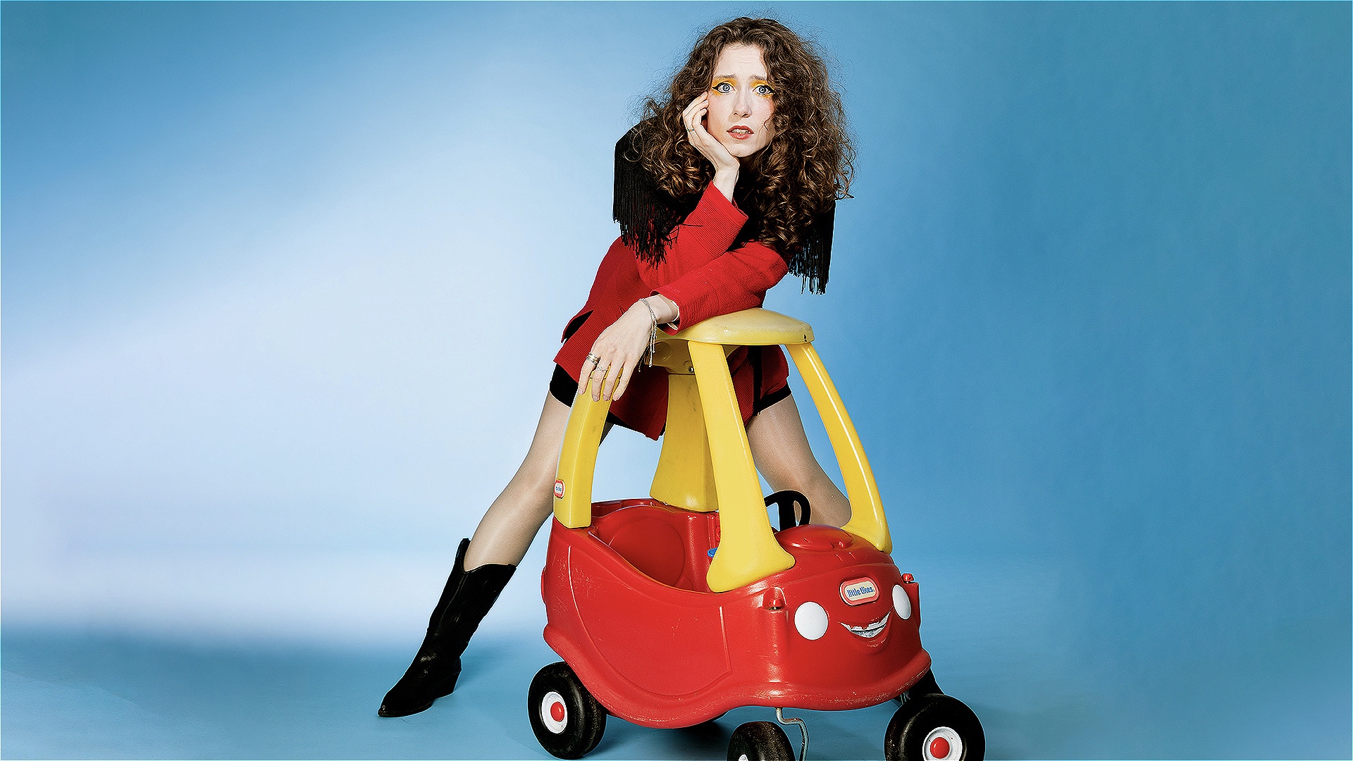 A woman leaning against a kids toy car, she is wearing long black boots and has brown curly hair. 
