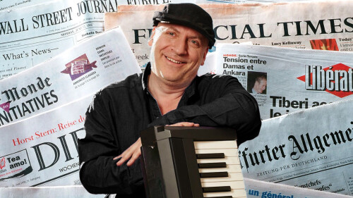 A man in the middle of the image is smiling and holding onto a keyboard. Behind him is a backdrop of newpapers. 