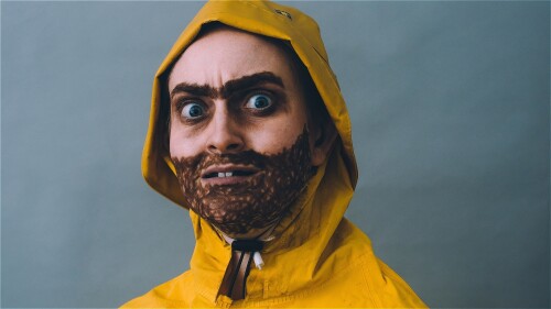 A man with a drawn on monobrow and beards wearing a yellow fishermans Sou'wester stares down the camera.