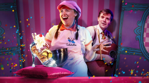 A performer in a pink apron and hat holding a wooden spoon. Behind her is a man wearing a pink apron and holding a guitar. 