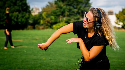 A woman with long blonde hair is wearing headphones and speaking through an attached headphone mic. She is mid dance. 