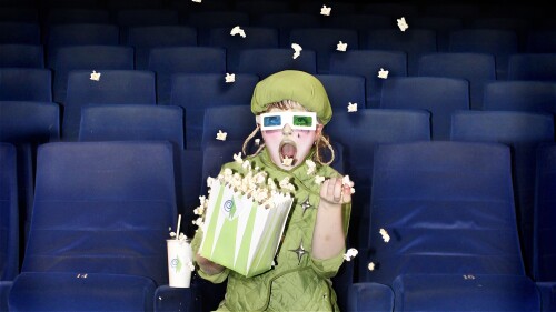 A woman in a green outfit is sat in the cinema holding a bag of popcorn. Kernals are flying everywhere and she is wearing 3D cinema glasses