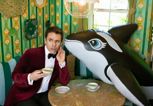 A man in a velvet tuxedo listens intently to an inflatable orca while sipping tea in a retro dining nook. It's playful, strange, and visually bold.