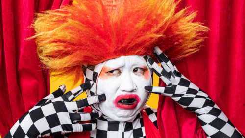 A person in white face make-up wearing a bright orange spiked wig, bright red lipstick and a black-and-white checkered-pattern morphsuit looks worried and holds both hands on their face in front of a red curtain background.