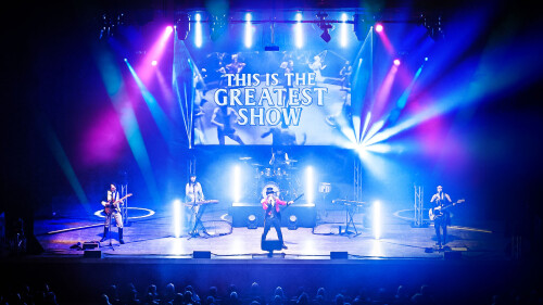 An image of a stage with 4 performers on holding instruments. Above is a projection which reads 'This is the greatest show' 