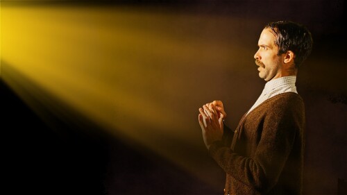 A man with a mustache and a brown cardigan stands in profile lit by a dramatic yellow beam, with his hands raised near his chest.