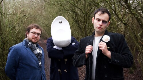 Two men stand in a forest, in between them is another person with a urinal on his head. They all look into the camera. 