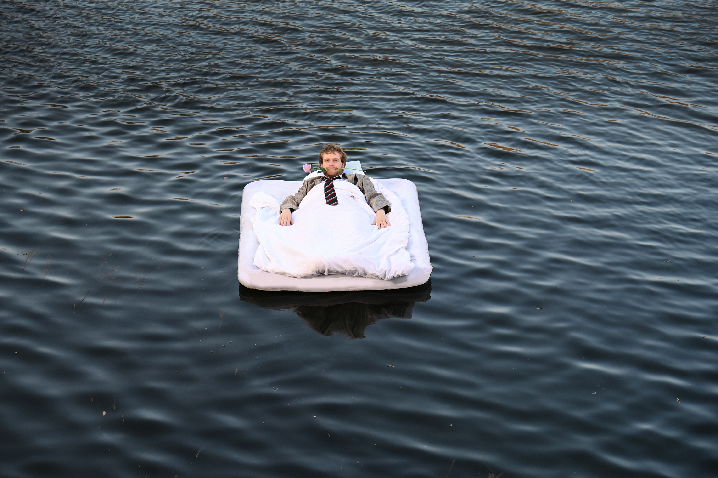 Theo lies on a mattress in a large body of water. He is wearing a suit and has a rose inbetween his teeth. 