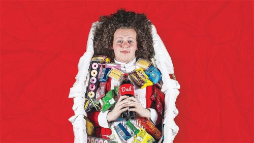 Jack Rooke lies in a coffin, he is covered in various snacks, sweets, crisps, biscuits and he holds a bottle of Coca Cola with 'Dad' written on it.
