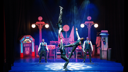 the black blues brothers on stage performing a stunt, behind them is a set of a smoky train station 